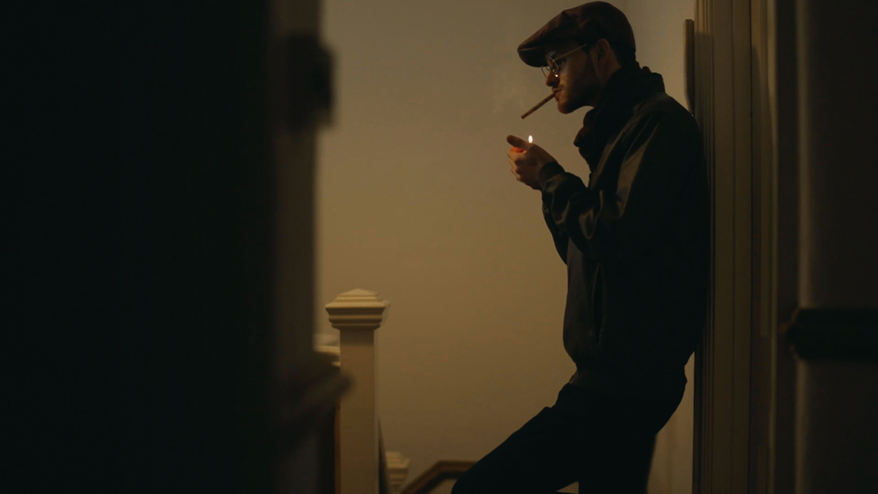 Still from a short film where Sam Hume leans on a wall, smoking